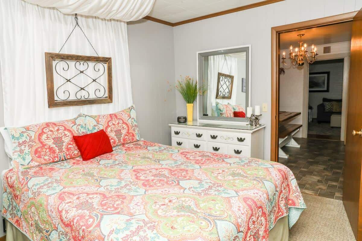 A bedroom with a large bed, a dresser with a mirror, a chandelier, and a door leading to another room.