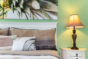 Een slaapkamer met een bed, kussens, een nachtkastje, een lamp en een plant aan de muur.
