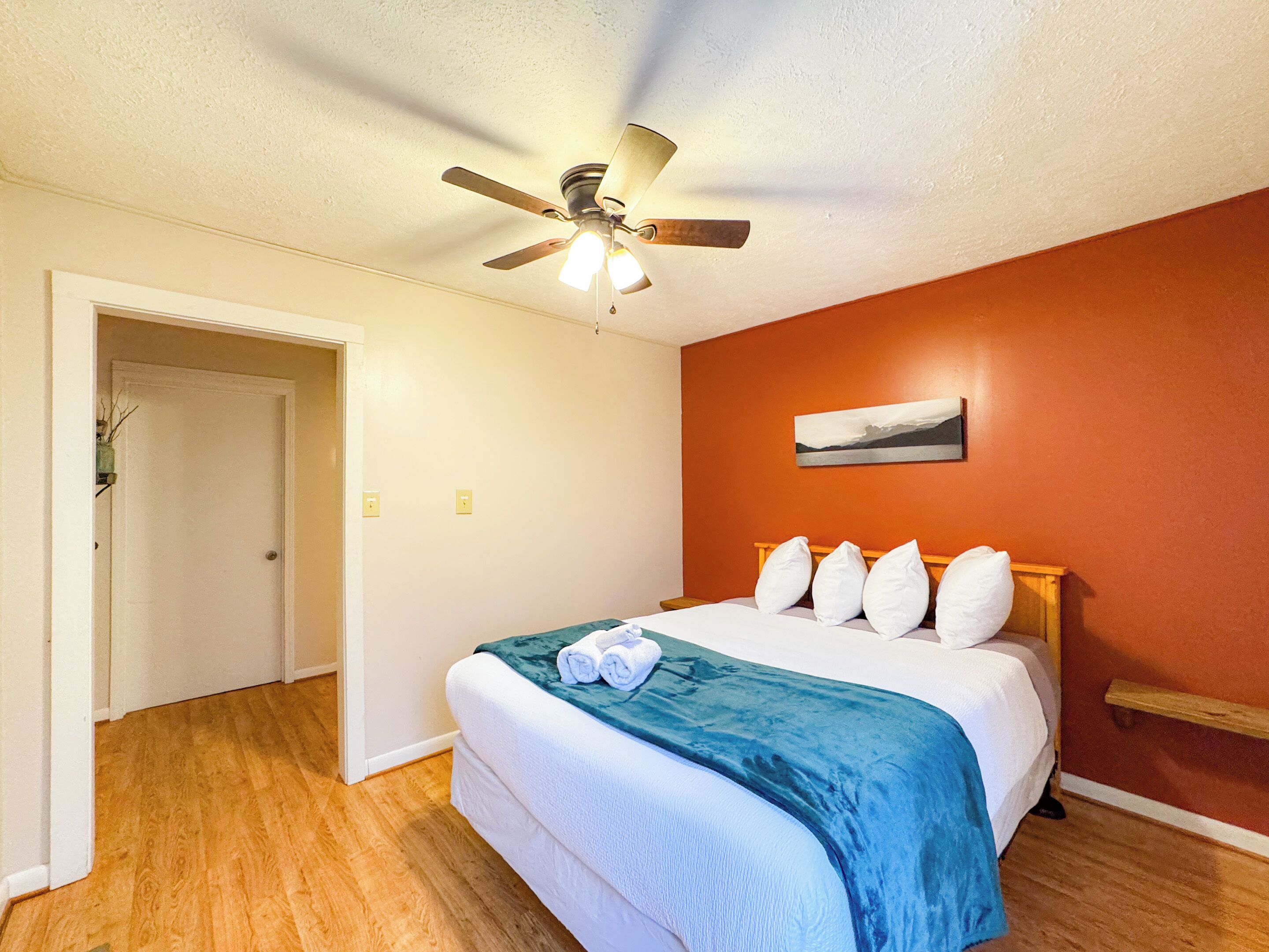 A hotel room with a bed, a ceiling fan, a painting on the wall, and wooden flooring.