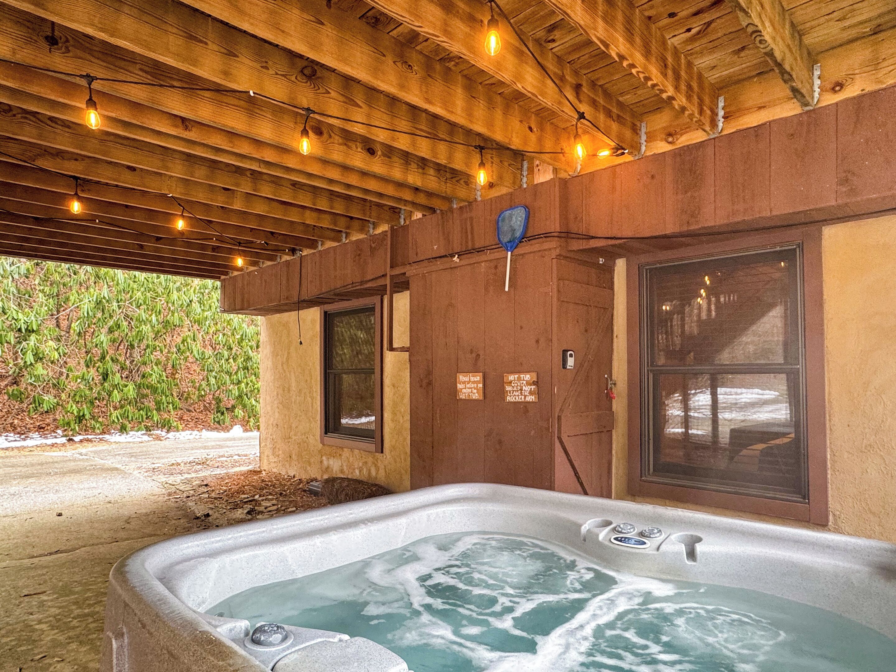 A hot tub under a wooden pergola with string lights, surrounded by a wooden cabin with large windows and a view of a green landscape.