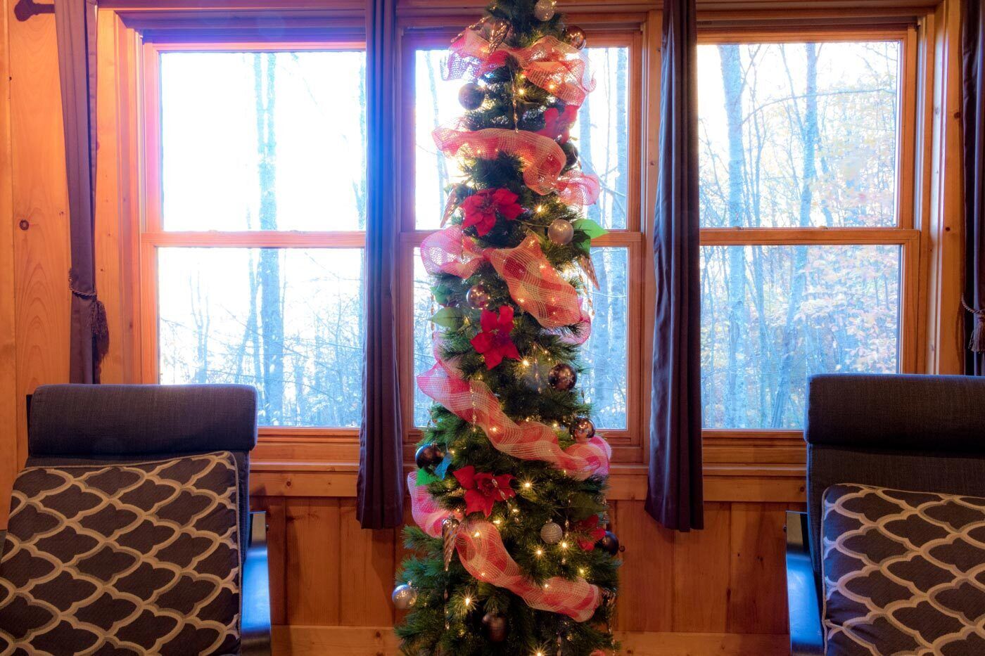 Ein gemütliches Zimmer mit Weihnachtsbaum, geschmückt mit Lichtern und Ornamenten, zwei gemusterten Sesseln und einem Fenster mit Blick auf Bäume.