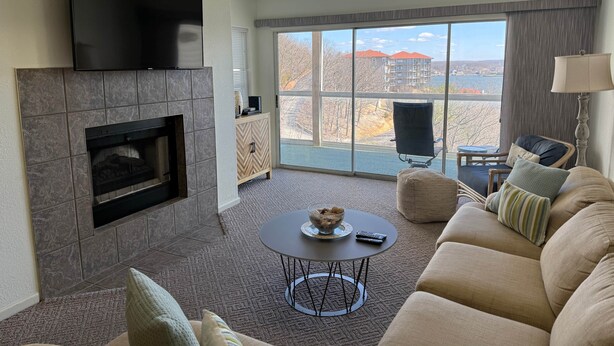 Ein Wohnzimmer mit Kamin, Flachbildfernseher, einer Couch, einem Couchtisch und Blick auf ein Gebäude sowie Bäume durch große Fenster.