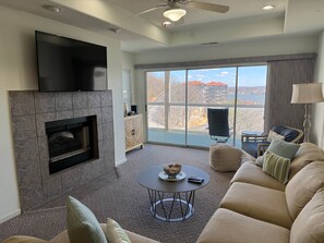 A living room with a fireplace, a flat-screen TV, a sofa, a coffee table, and a view of a building and trees through large windows. - Great view-Screened Deck, Must See! 'TopSider' 3-bed 3-bath (Lake Ozark)