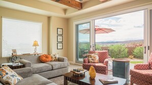 A spacious living room with a grey sofa, a coffee table, and a red patterned armchair. There is a large window with a view of a desert landscape. - Sun-e-Scape- Relax in the Desert! (Washington)