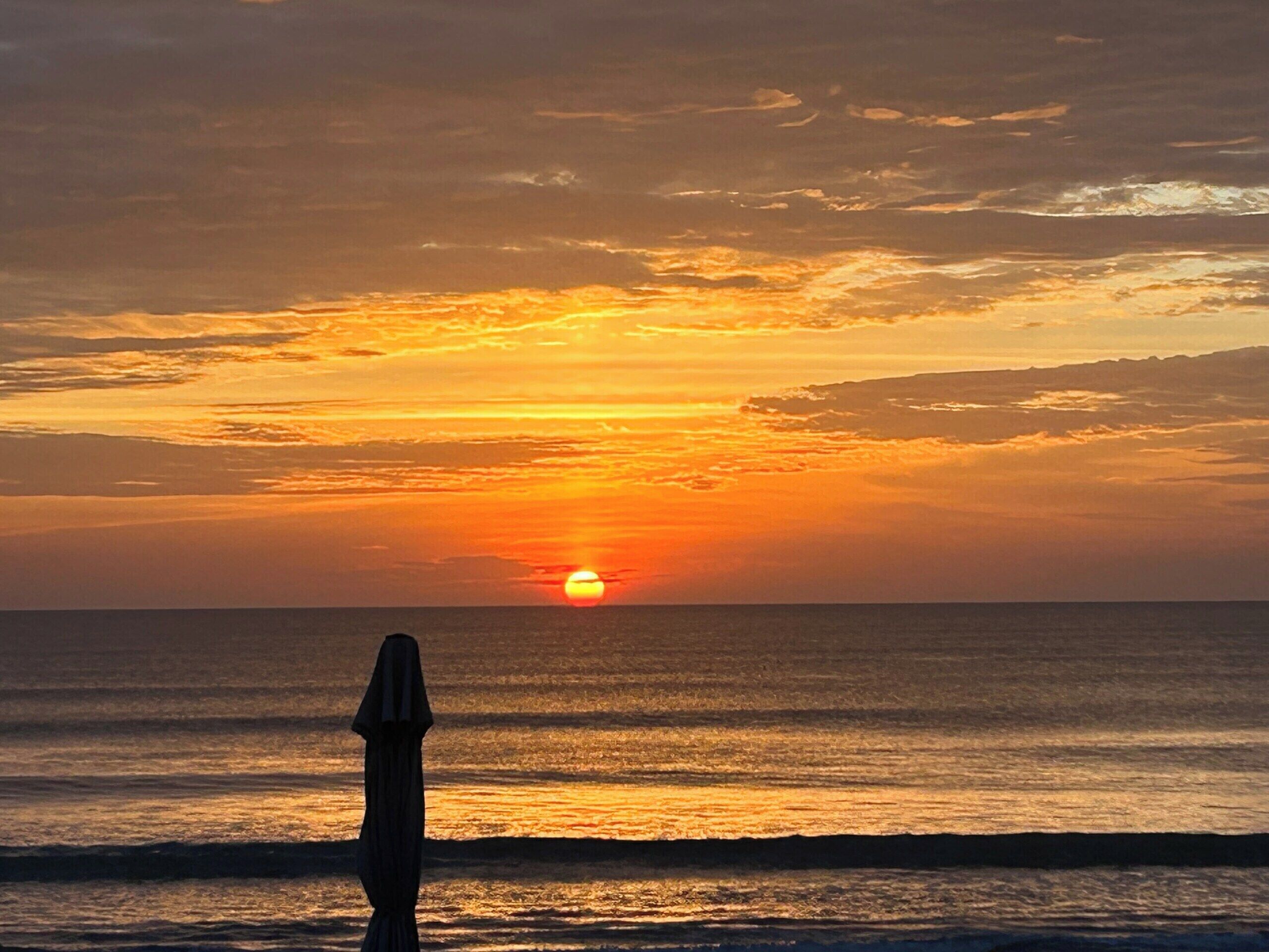 Ein Sonnenuntergang über dem Meer mit der Silhouette einer Person, die am Strand steht.