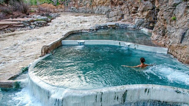 Eine natürliche heiße Quelle mit einer Person im Wasser, umgeben von felsigem Gelände und einem Wasserfall.