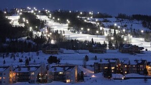 Un paysage enneigé avec des pistes de ski illuminées et des bâtiments au pied des montagnes, ce qui évoque un lieu de villégiature hivernale.
