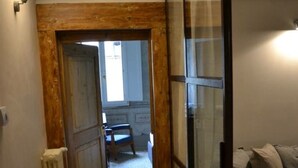 Een smalle gang met houten vloer en een radiator aan de linkerkant. Een deur leidt naar een kamer met een bed en een raam.
