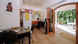 Une cuisine compacte comprenant un coin repas, un réfrigérateur, une cuisinière et une vue sur le jardin grâce à une porte ouverte.