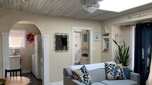 A cozy living room with a grey sofa, wooden flooring, a chandelier, and a view of the kitchen through an archway.