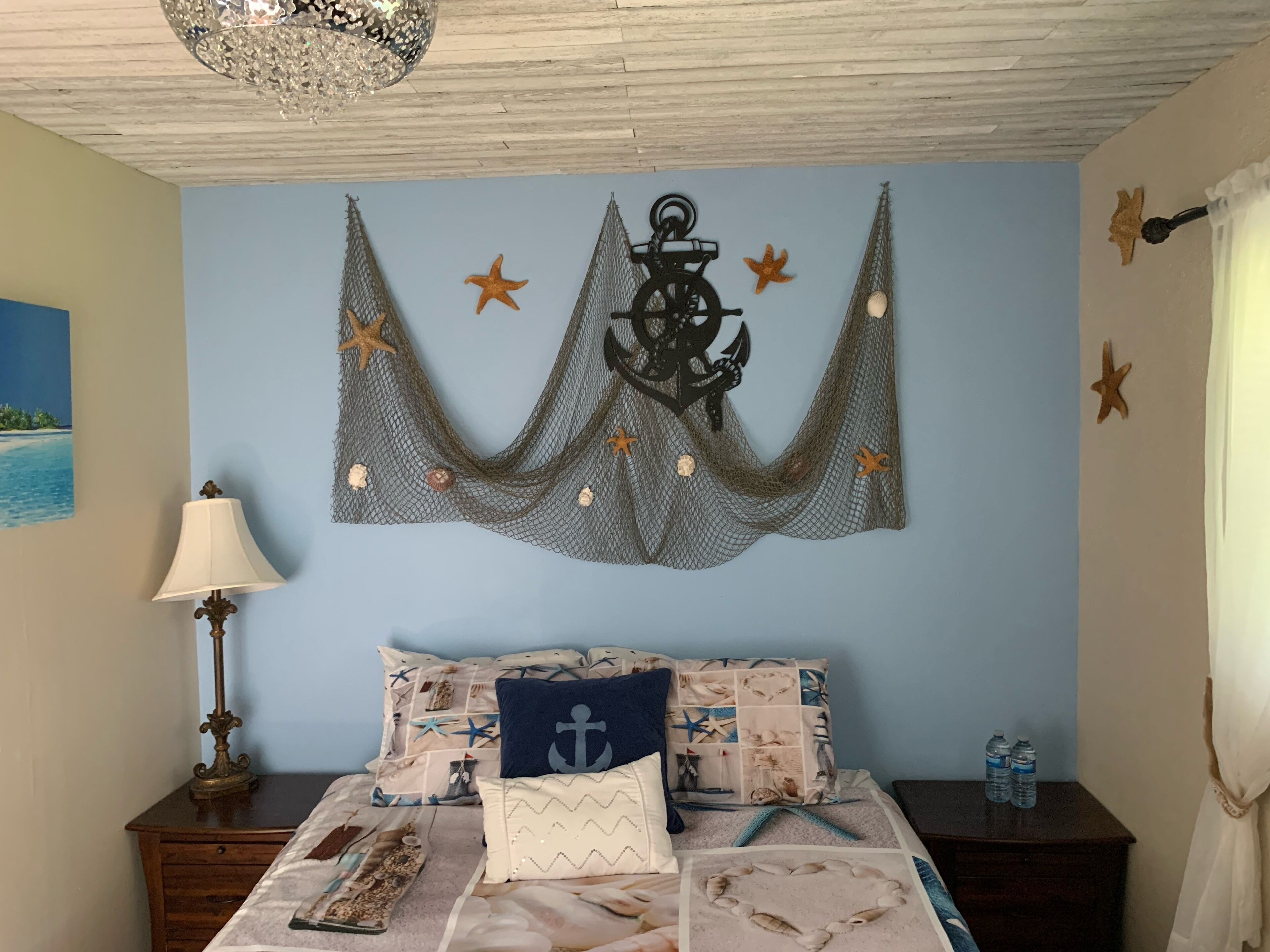 A bedroom with a nautical theme, featuring a bed with starfish and anchor decorations, a lamp, and a painting of a beach.