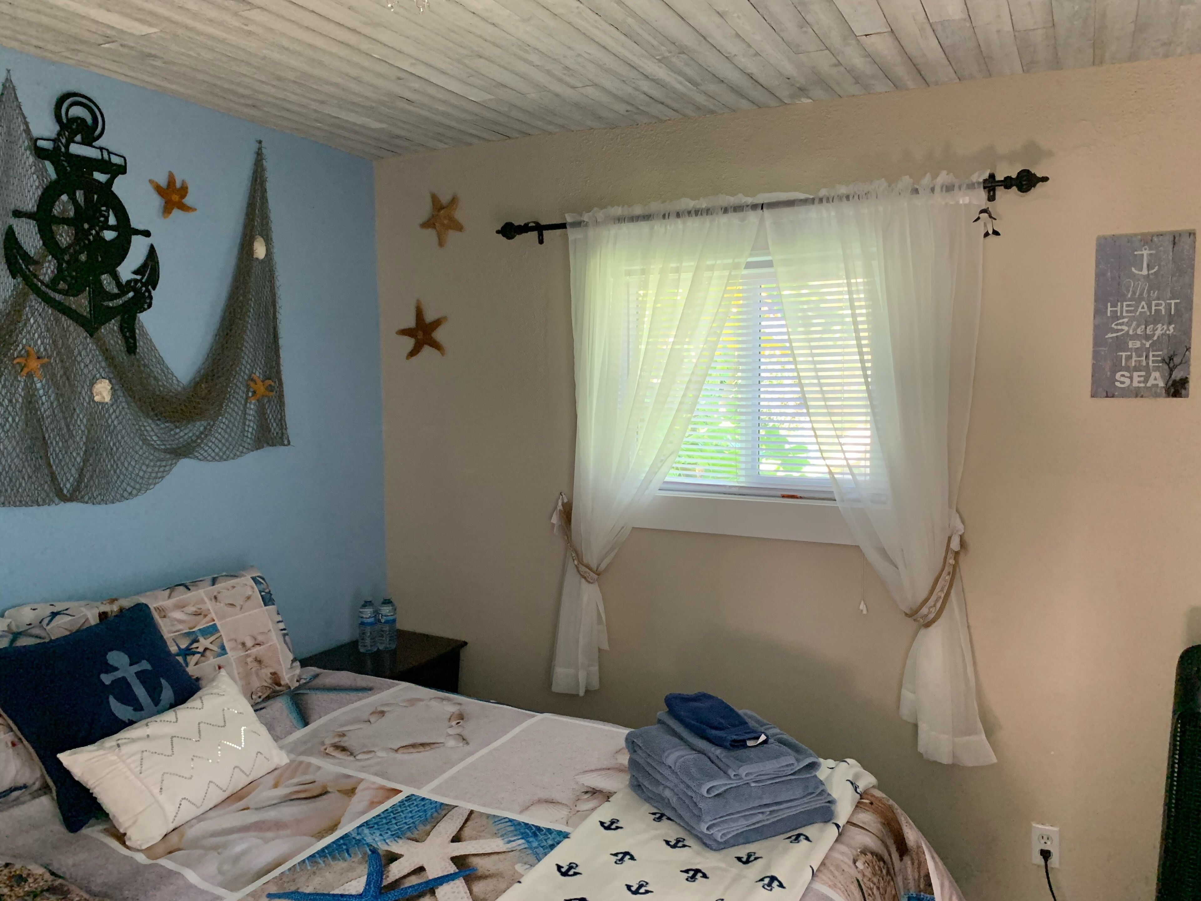 A bedroom with a bed, a window with sheer curtains, starfish decorations, and a wall with nautical-themed decor.