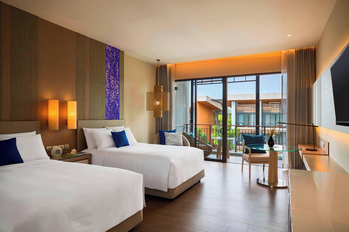A hotel room with two beds, a glass table, and a view of the outdoors.