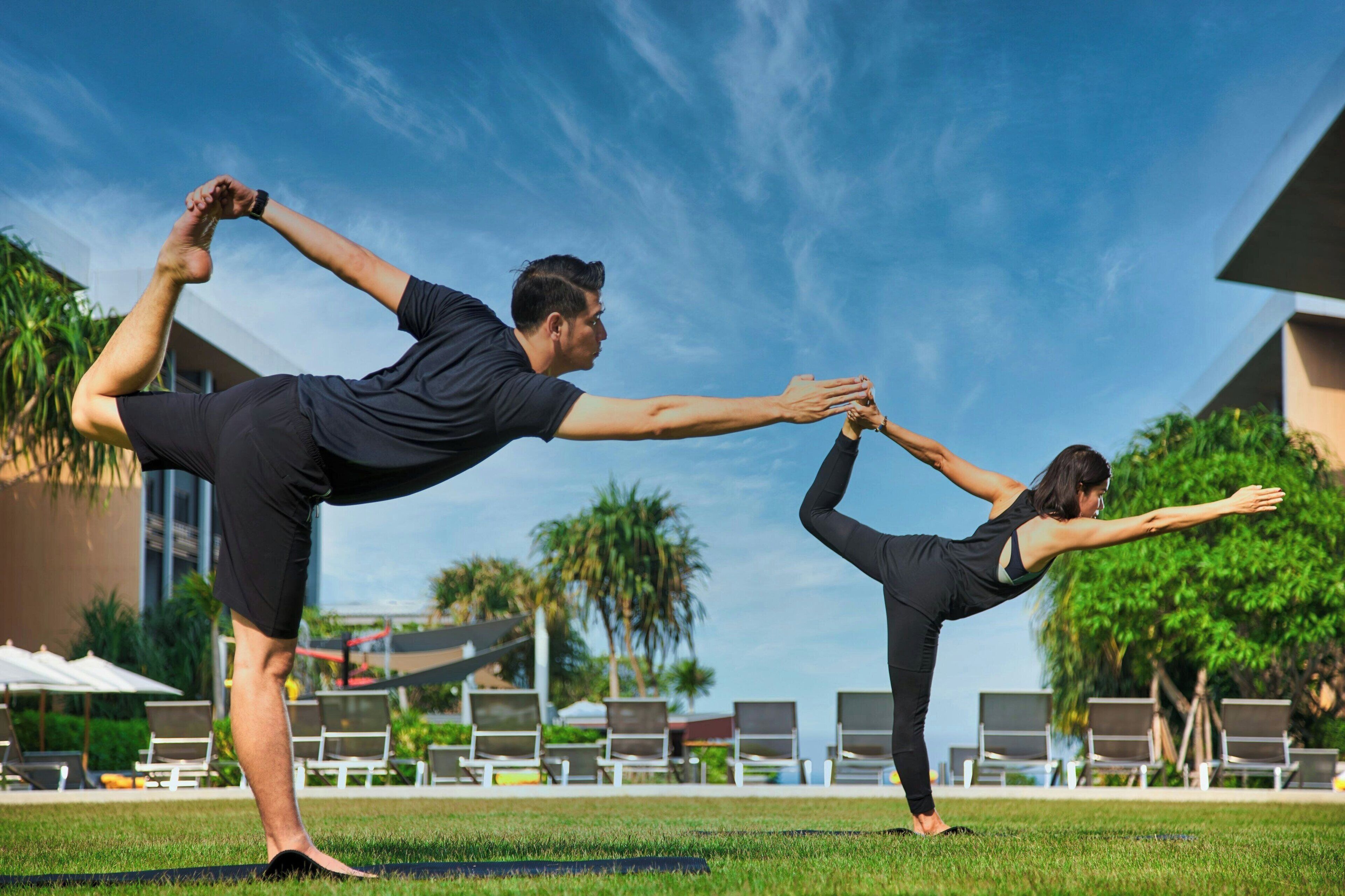 Two people practicing yoga outdoors with a pool and lounge chairs in the background.