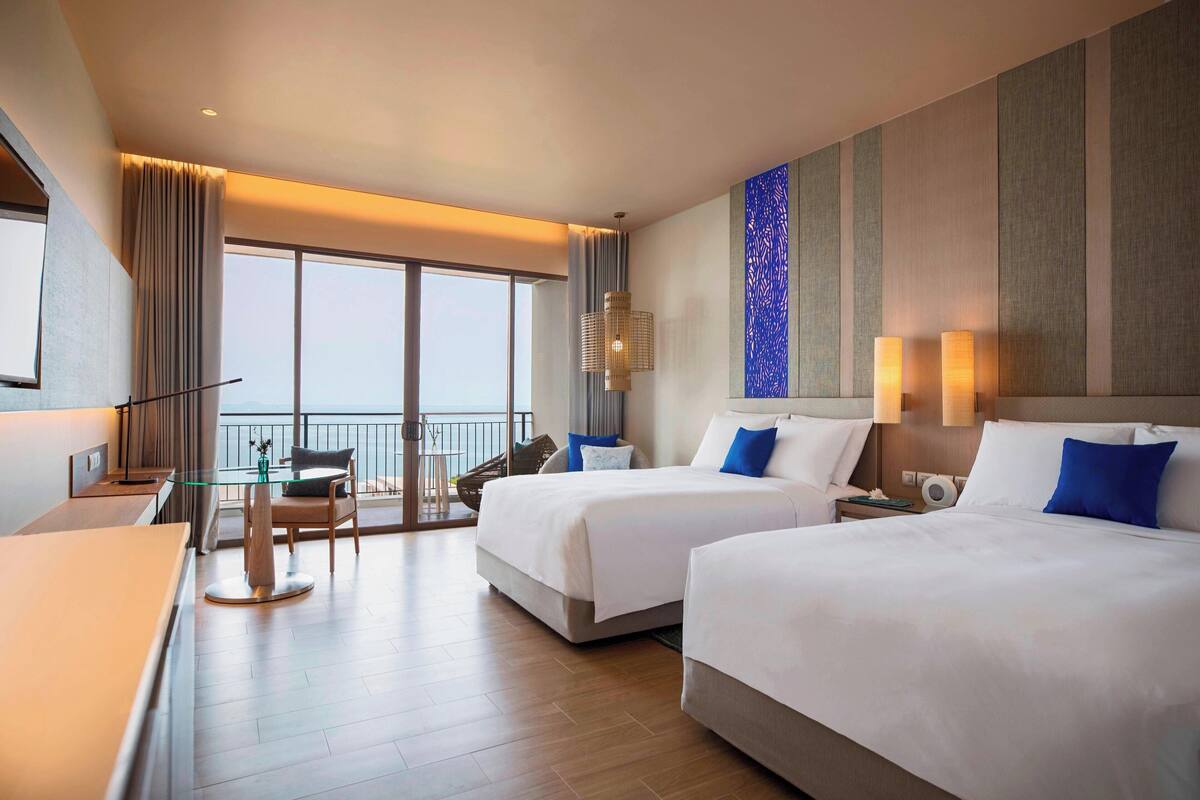 A hotel room with two beds, a dining table, chairs, a balcony with a view, and a large window.