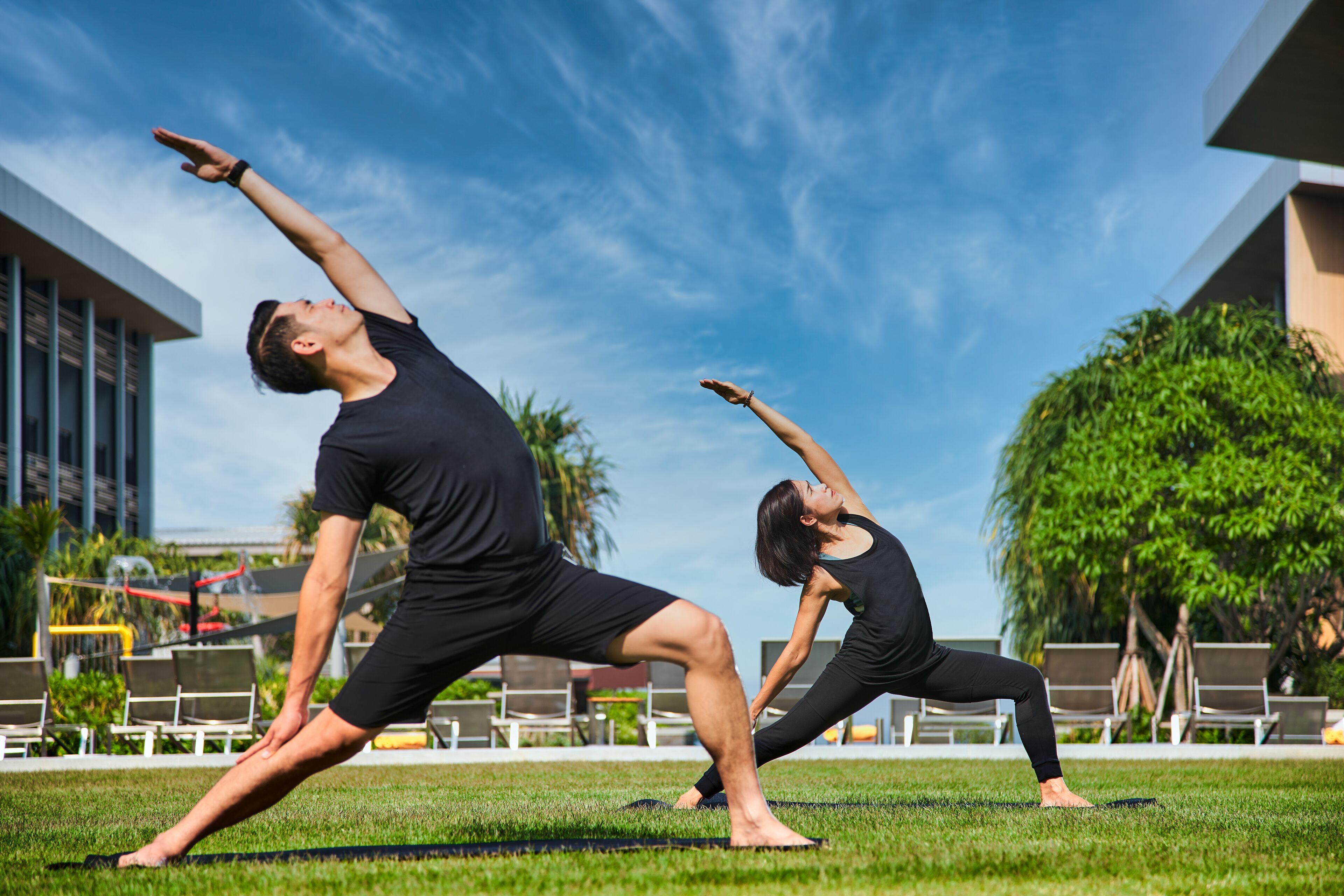 Two people performing a yoga pose on a grassy area with a building and chairs in the background.