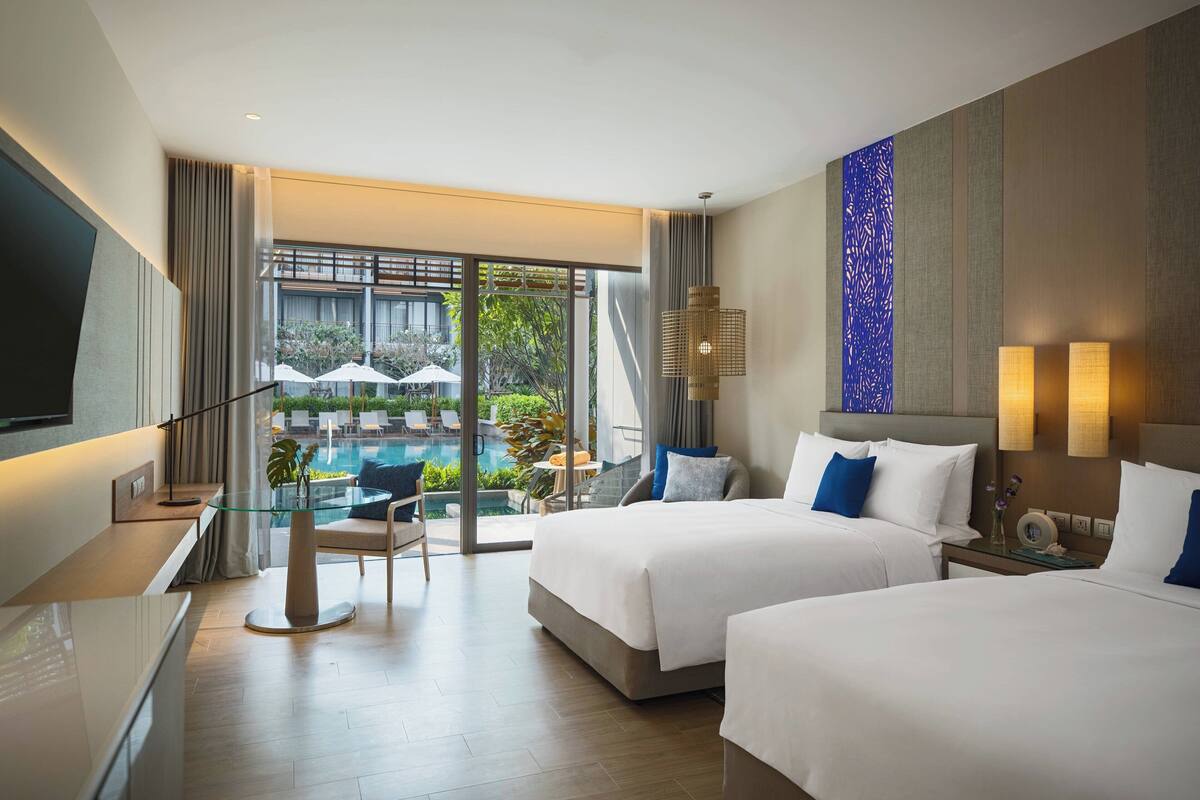 A hotel room with two beds, a glass table, and a view of an outdoor pool area.