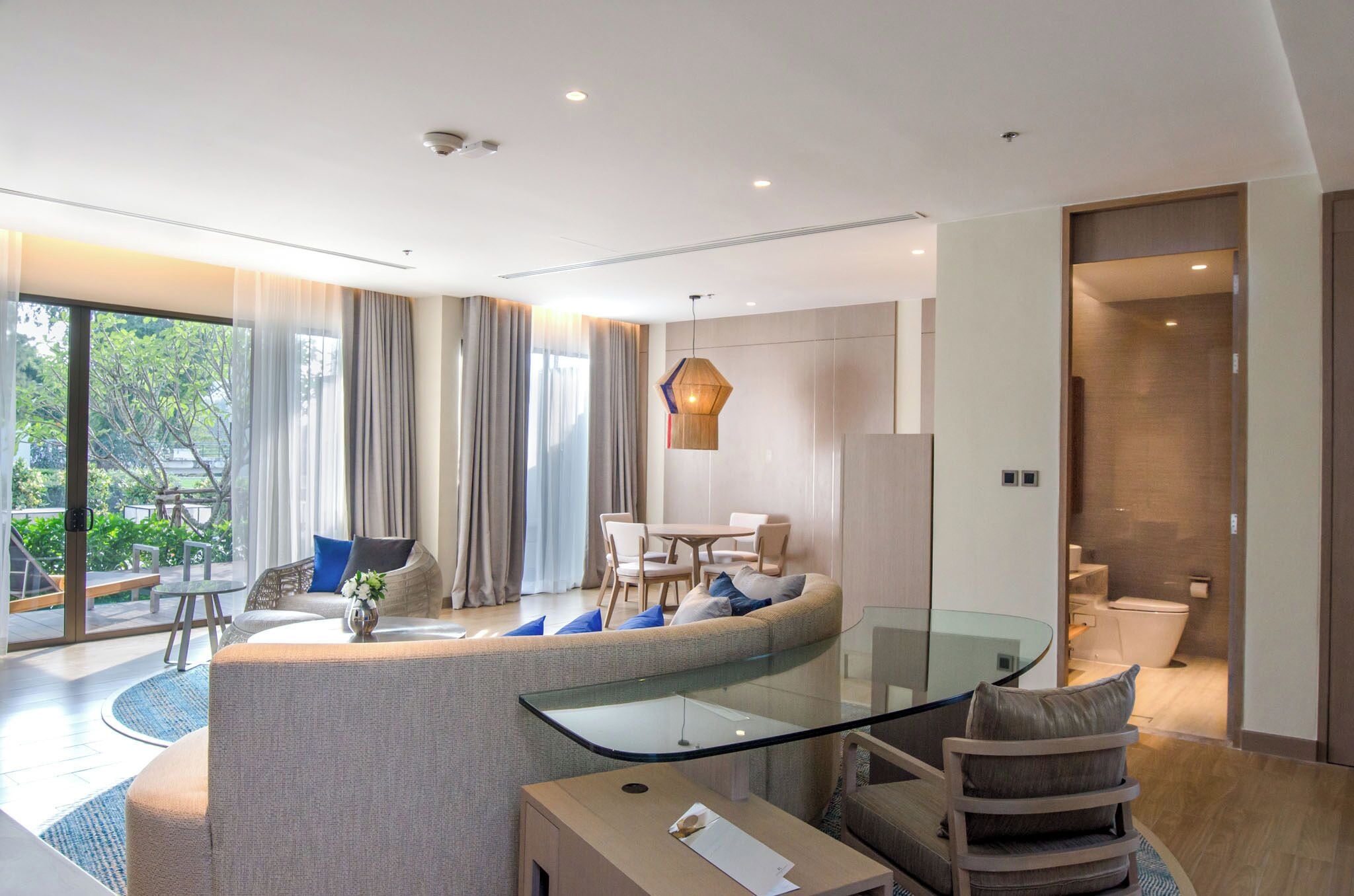 Una habitación de hotel moderna con un sofá curvo, una mesa de centro de vidrio, una zona de comedor con mesa y sillas, y un baño a la vista.