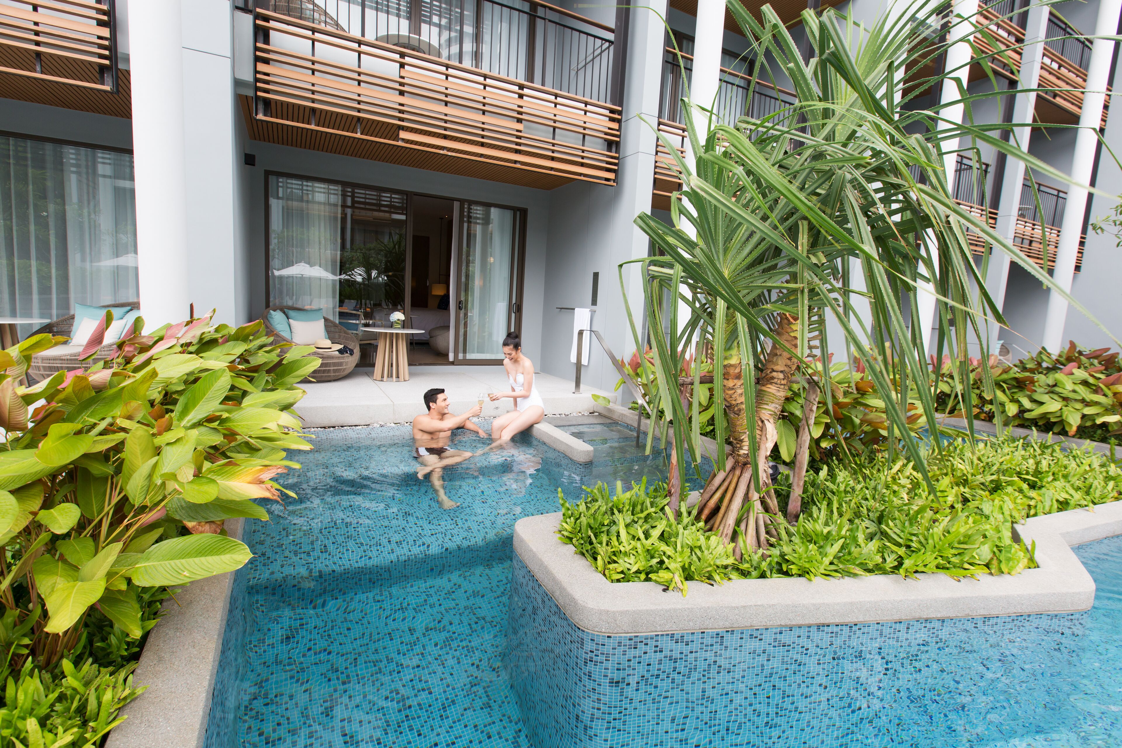 Zona de piscina con dos personas relajándose, rodeada de vegetación exuberante y un edificio moderno con balcones.