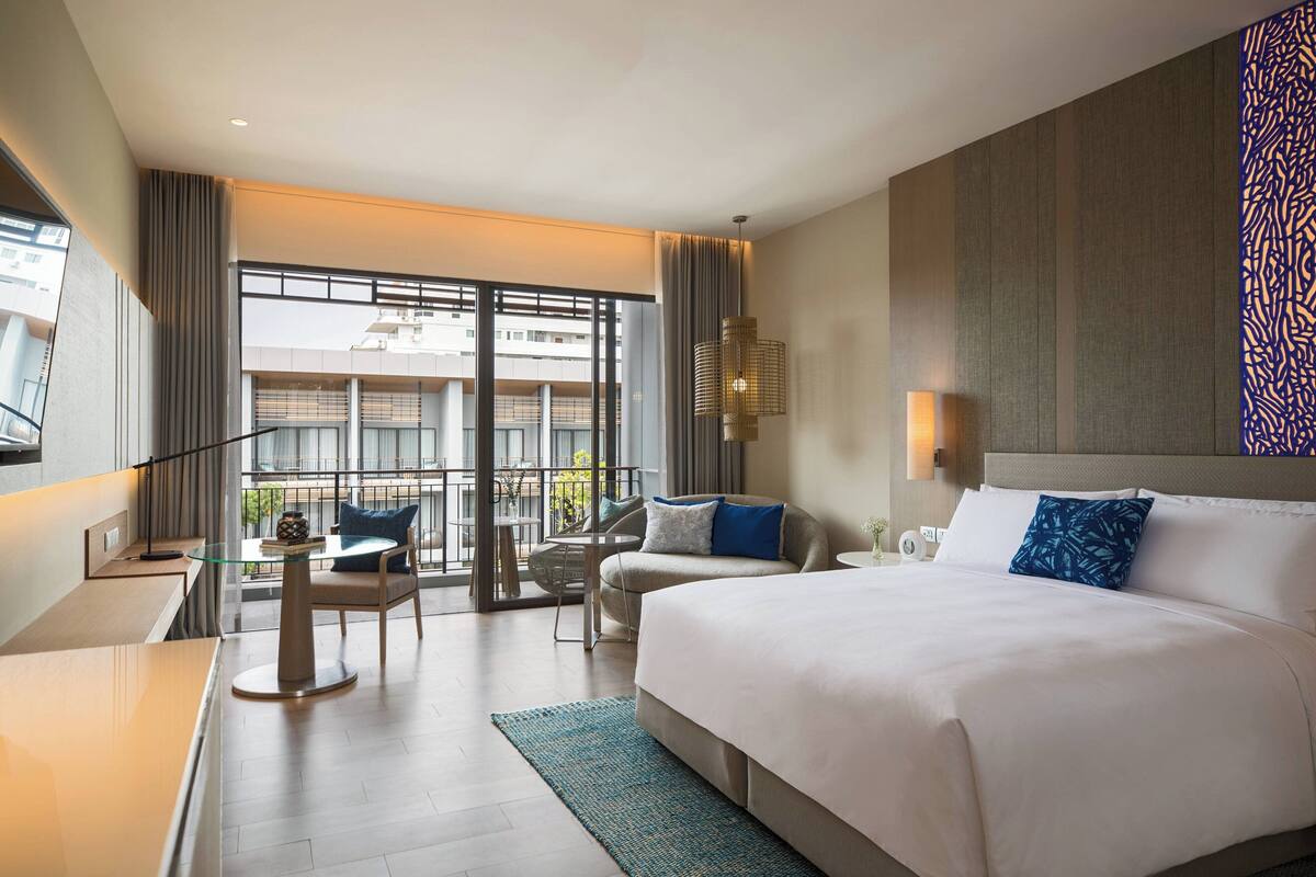A modern hotel room with a large bed, a desk, a chair, and a balcony with a view of the city.