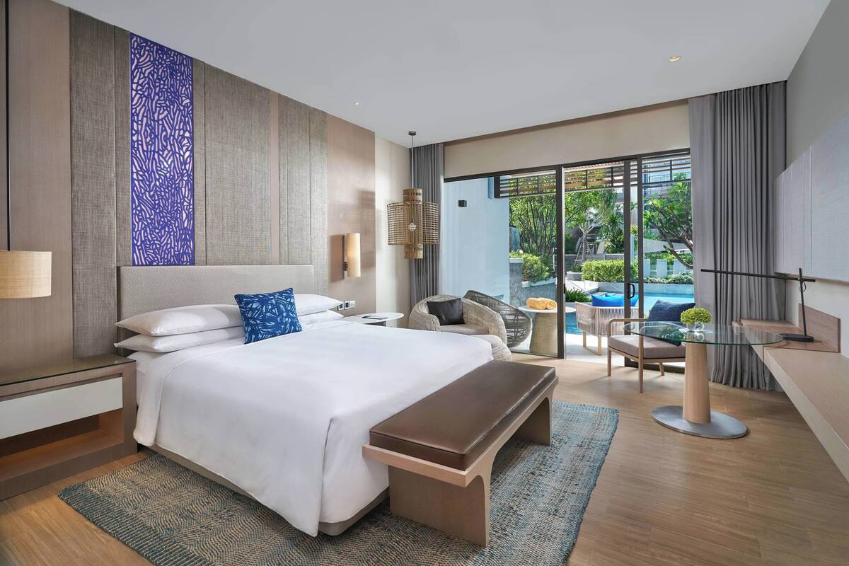 A modern hotel room with a large bed, a bench, a glass table, and a view of the outdoors.