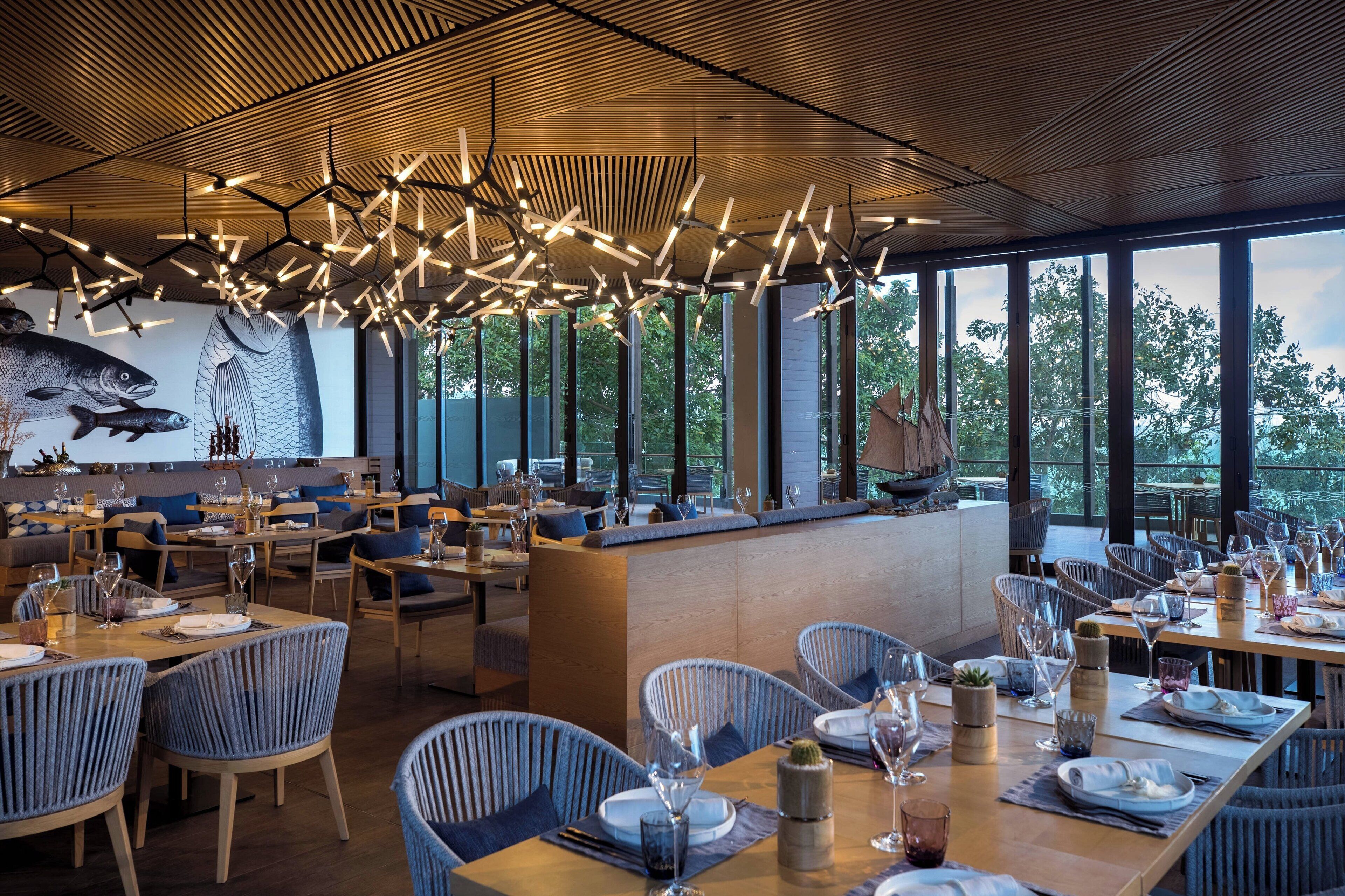 Un restaurante moderno con techos altos, luminarias únicas y grandes ventanales que ofrecen vistas al exterior.