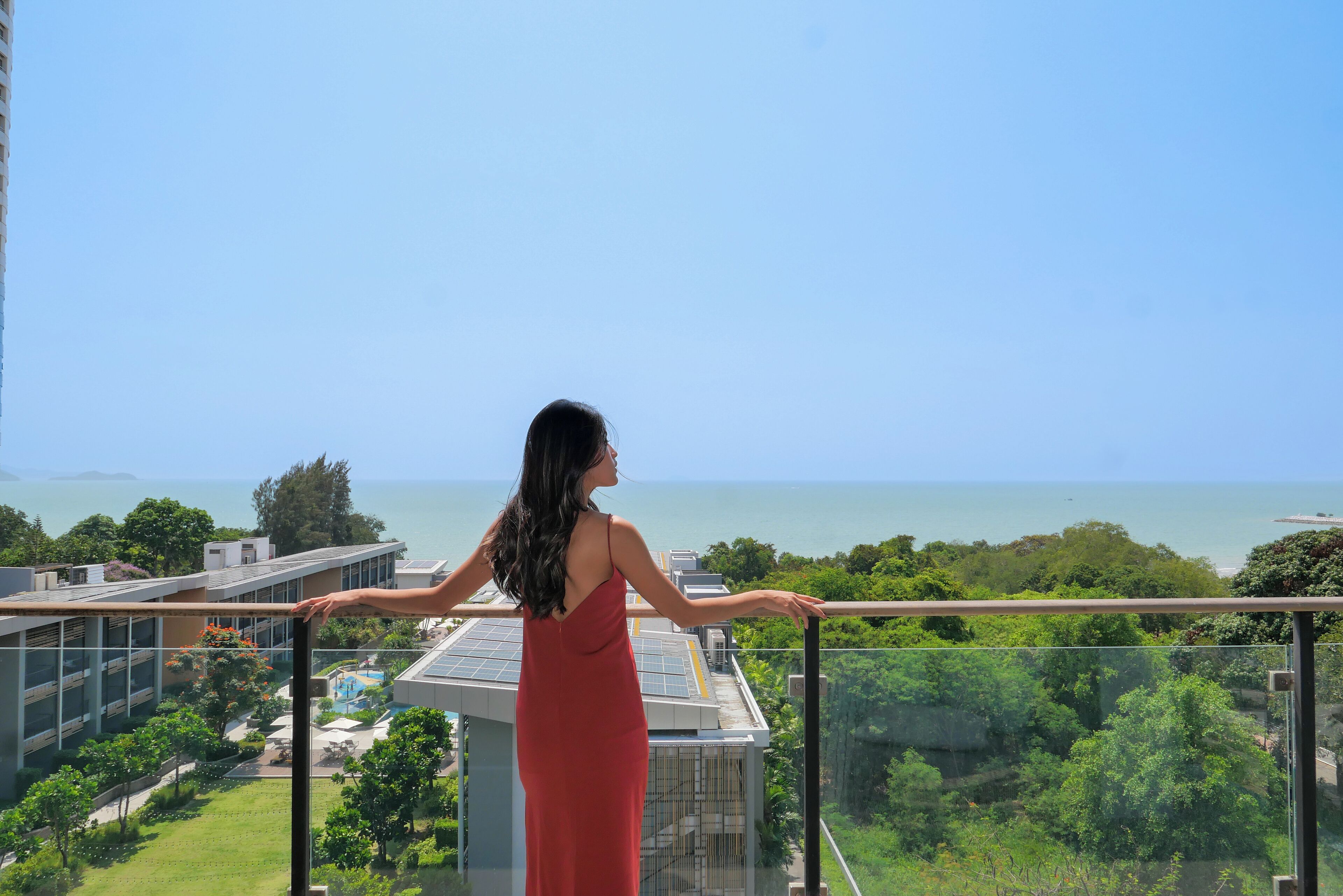 A woman in a red dress standing on a balcony with a view of a pool, greenery, and a body of water.