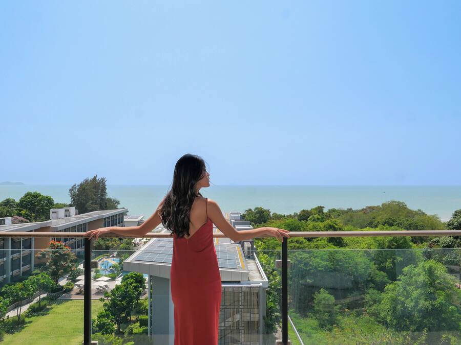 A woman in a red dress standing on a balcony with a view of a pool, greenery, and a body of water.