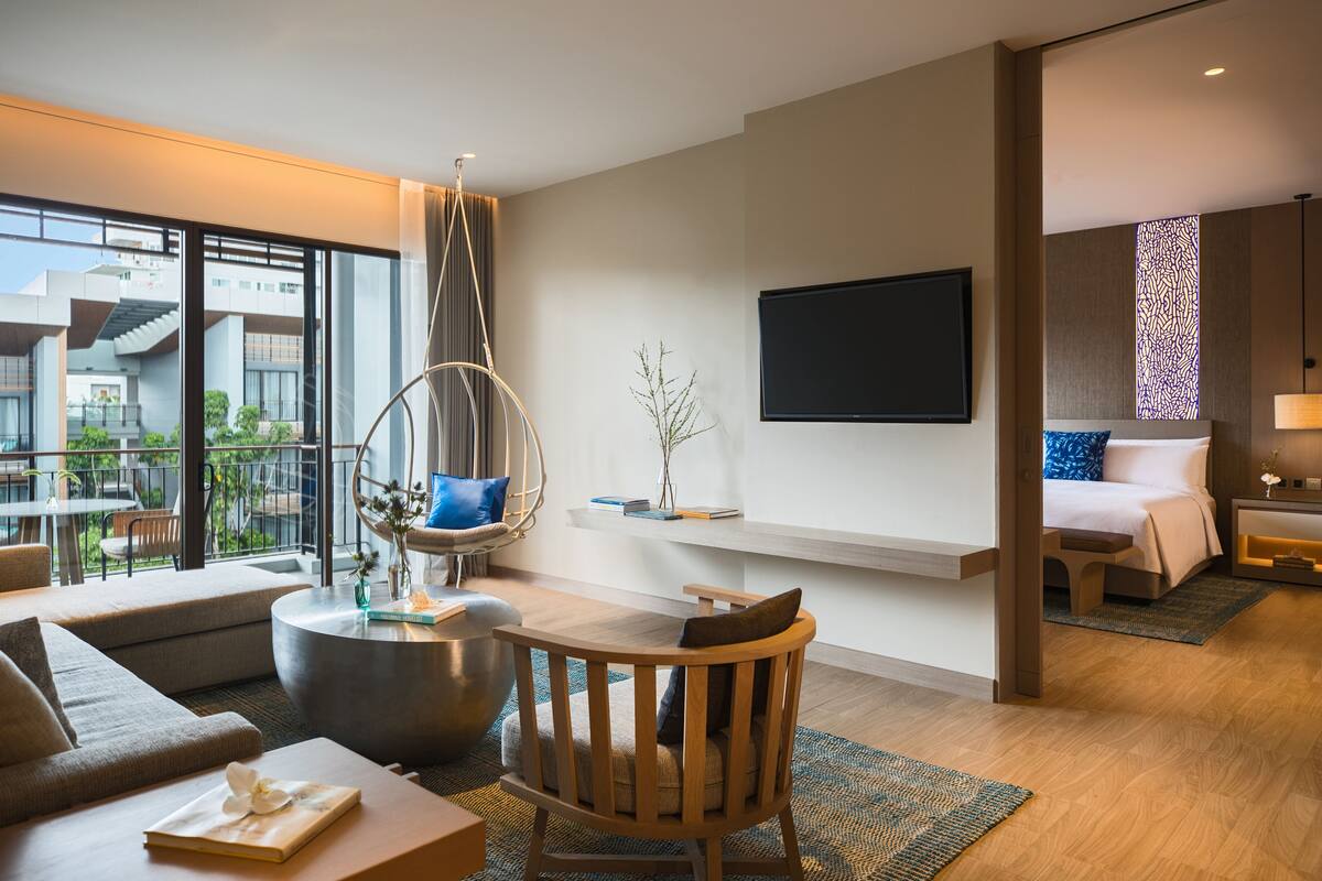 A modern hotel room with a large window, a hanging chair, a flat-screen TV, a bed with a patterned headboard, and a small table with books and a vase.