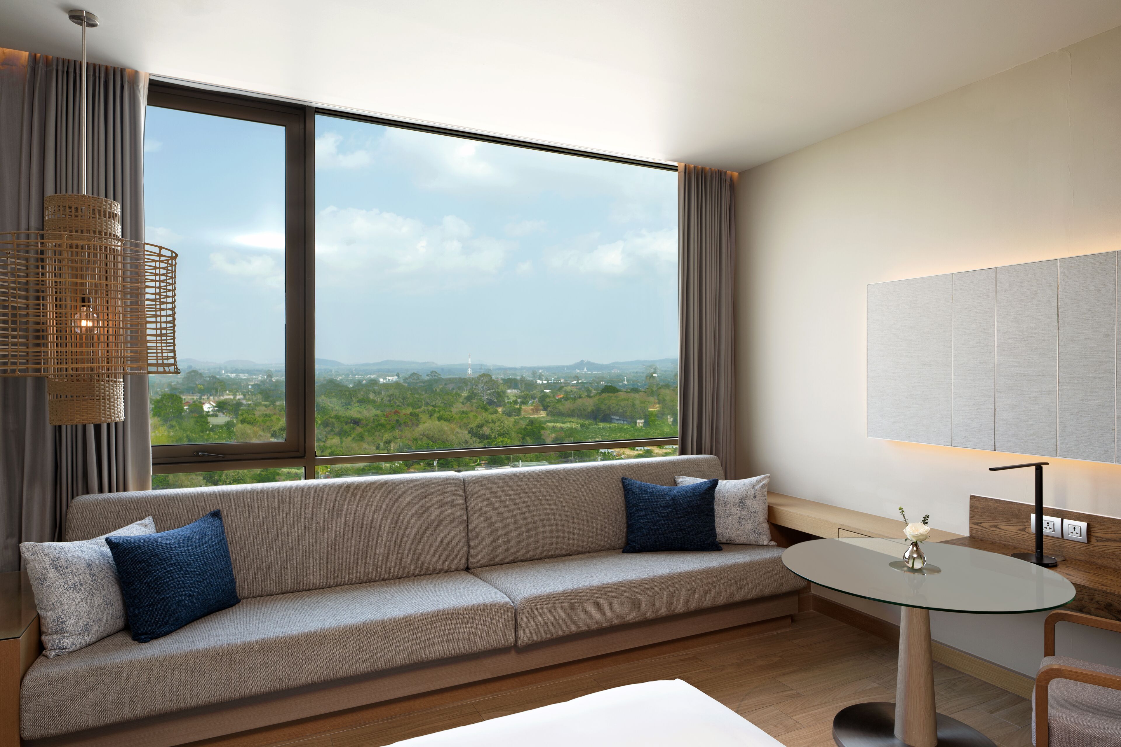 A modern hotel room with a large window offering a city view, a sofa with blue cushions, a small round table, and a hanging light fixture.