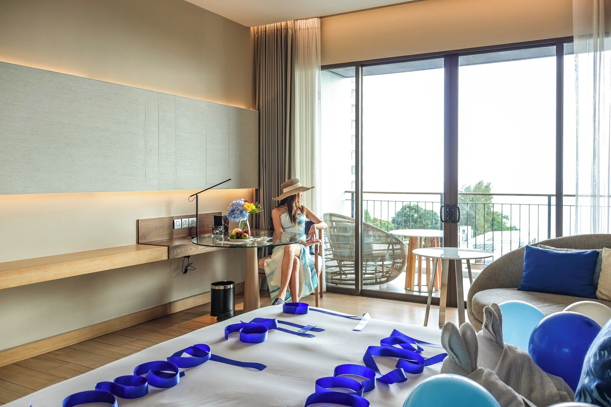 A hotel room with a balcony, a desk with a laptop, a bed with blue ribbons, and a sofa.