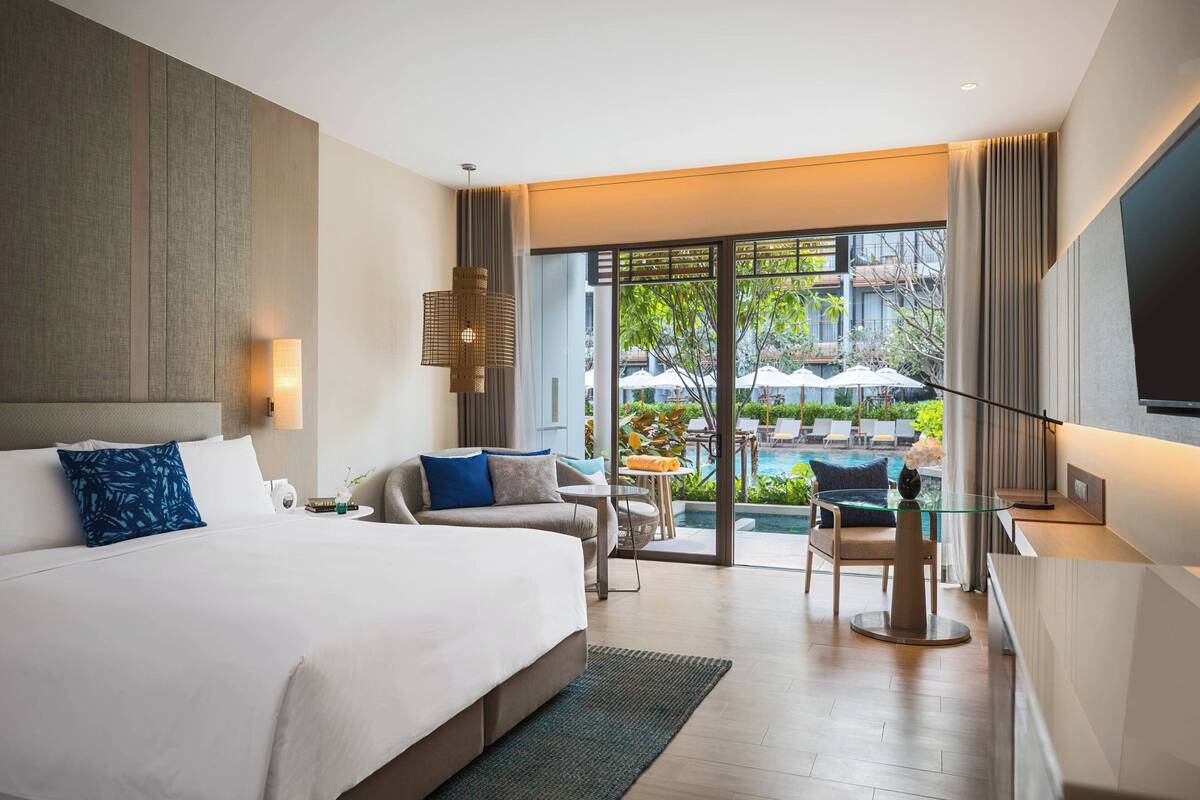 A modern hotel room with a large bed, a glass table, and a view of an outdoor pool area.