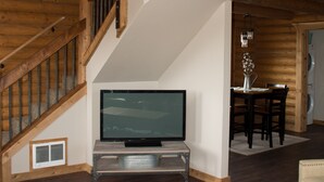 A spacious living area with a wooden staircase, a flat-screen TV, a dining table, and a sofa.