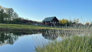 A tranquil lakeside scene with a wooden cabin, clear skies, and surrounding greenery.