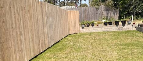 A fenced backyard with a grassy area, potted plants, and trees in the background.