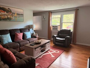 A living room with a brown leather sofa, a coffee table, a red rug with a white design, a window with curtains, and a painting on the wall. - New 2 bedroom Cape Cod Vacation Rental 9 mins to the beach! (Yarmouth)