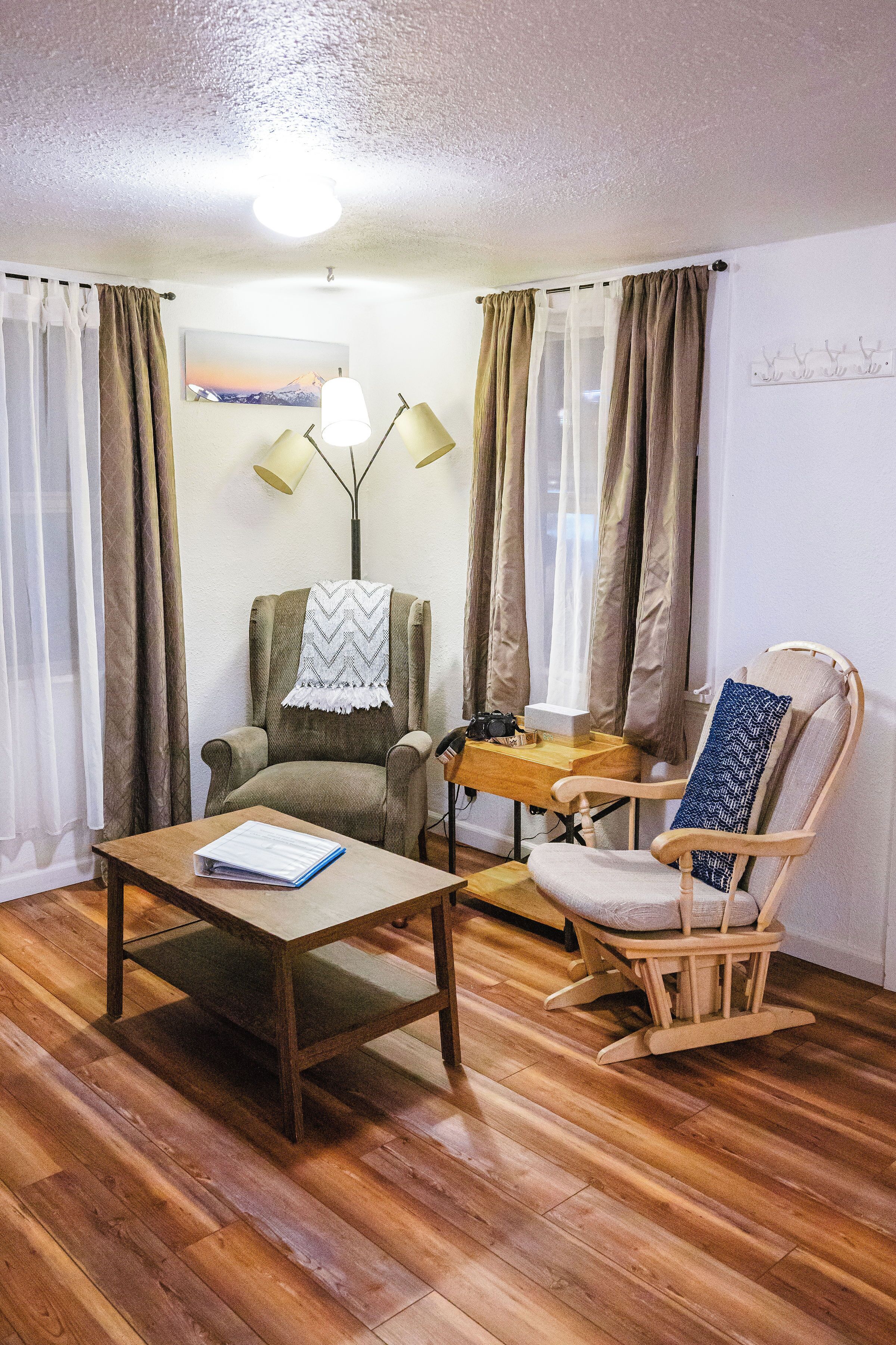 A room with a wooden floor, a green armchair with a blanket, a wooden table with a book, a wooden rocking chair, and a lamp.