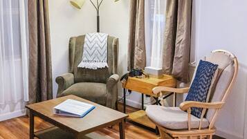 A room with a wooden floor, a green armchair with a blanket, a wooden table with a book, a wooden rocking chair, and a lamp.