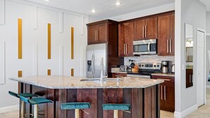 A kitchen with dark wooden cabinets, a central island with stools, stainless steel appliances, and a white backsplash. - Comfortable 4bdr w/private pool - No resort fees (Kissimmee)