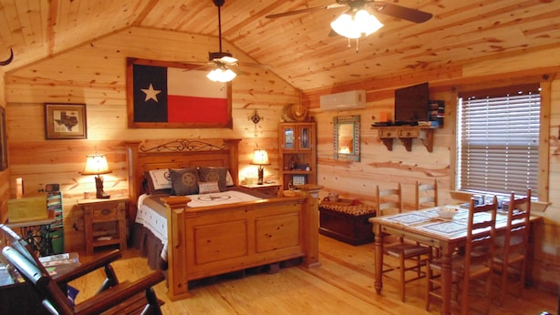 A cozy log cabin room with a bed, a dining table with chairs, a ceiling fan, and a Texas flag. - Texas T Bed and Breakfasts (Llano)