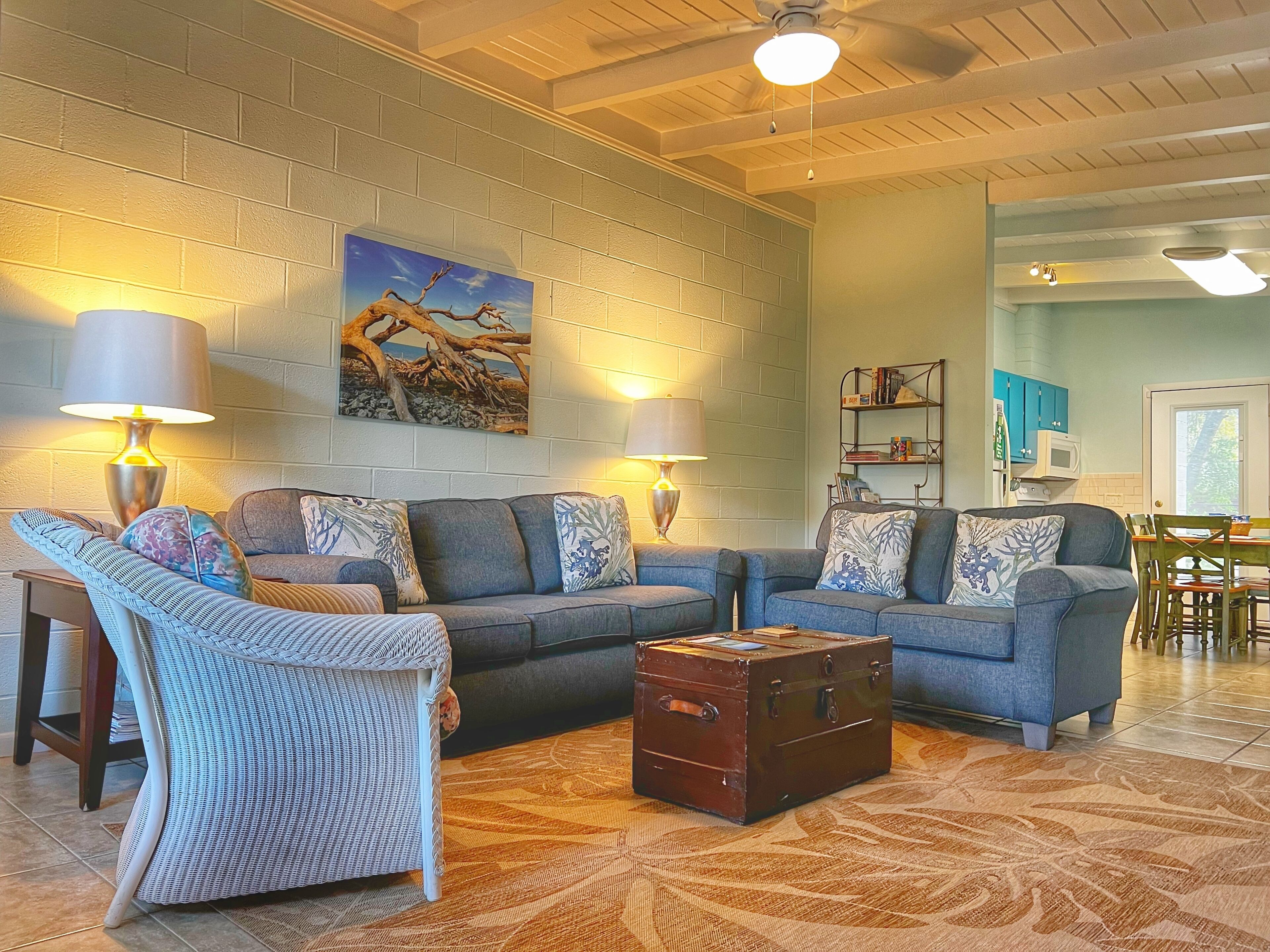 A living room with a sectional sofa, two floor lamps, a painting, a wooden chest, and a dining area in the background.