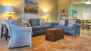 A living room with a sectional sofa, two floor lamps, a painting, a wooden chest, and a dining area in the background. - A Beach, Golf, Tennis and Fun for the whole family Sandy Toes & Salty Kisses A (Jekyll Island)