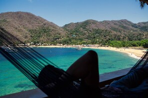 Blick auf einen Strand mit klarem türkisblauem Wasser, einer Hängematte und einem Bergpanorama im Hintergrund.
