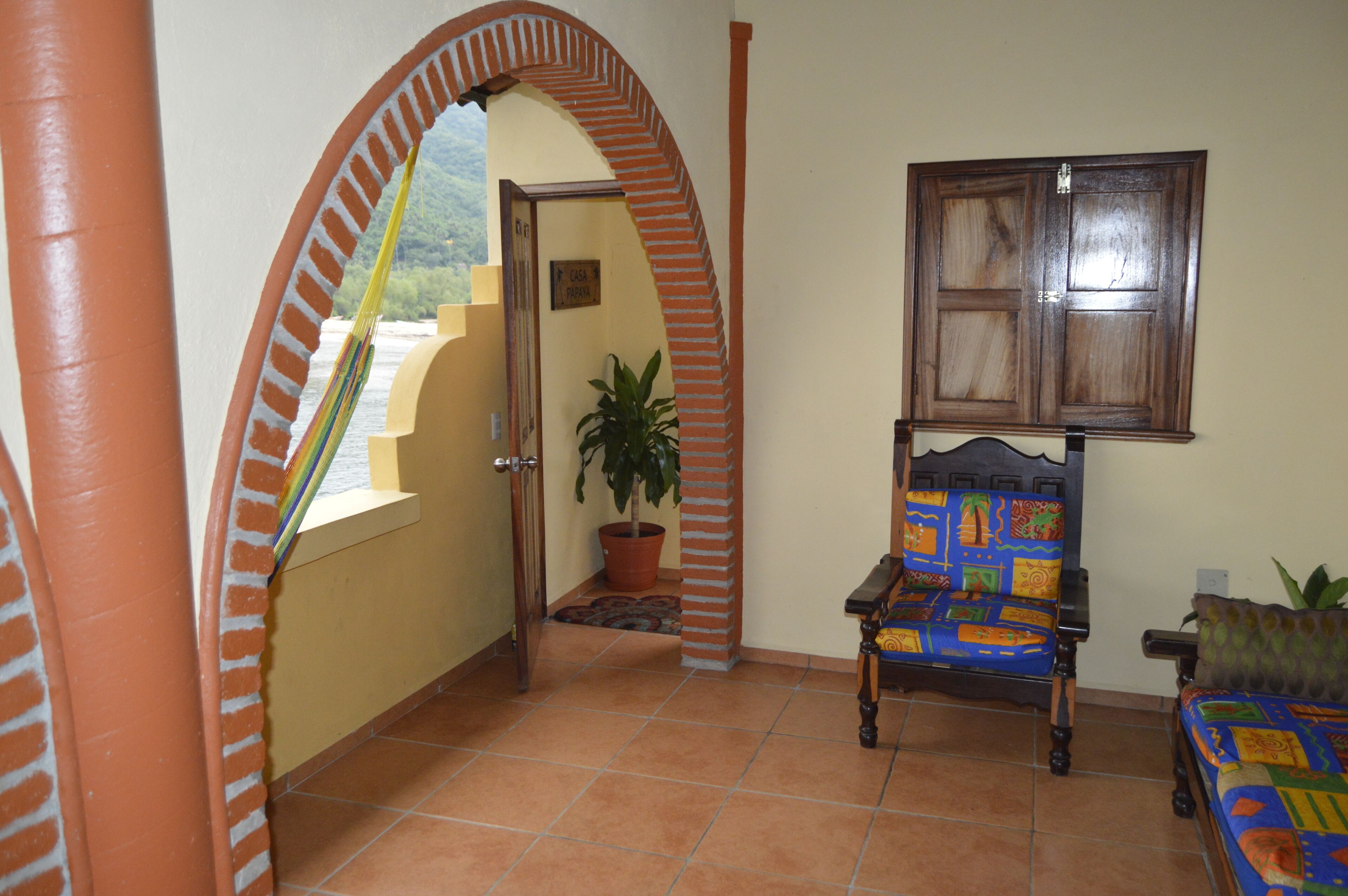 A room with a tiled floor, a wooden chair with a colorful cushion, a potted plant, a wooden door, and a window with a wooden frame.
