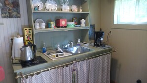 Une cuisine équipée d’un évier, d’une bouilloire et d’une machine à café. Au-dessus de l’évier, on trouve des étagères où sont rangés de la vaisselle, ainsi qu’une horloge.