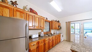 A kitchen with wooden cabinets, a stainless steel refrigerator, a granite countertop, and a view of the ocean through a sliding glass door. - Beach House Condos: Beachfront Ocean House - 7mile Beach Jamaica (Negril)