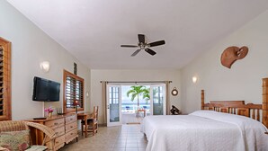 A bedroom with a bed, a desk, a chair, a television, and a view of the beach through an open door. - Beach House Condos: Beachfront Ocean House - 7mile Beach Jamaica (Negril)