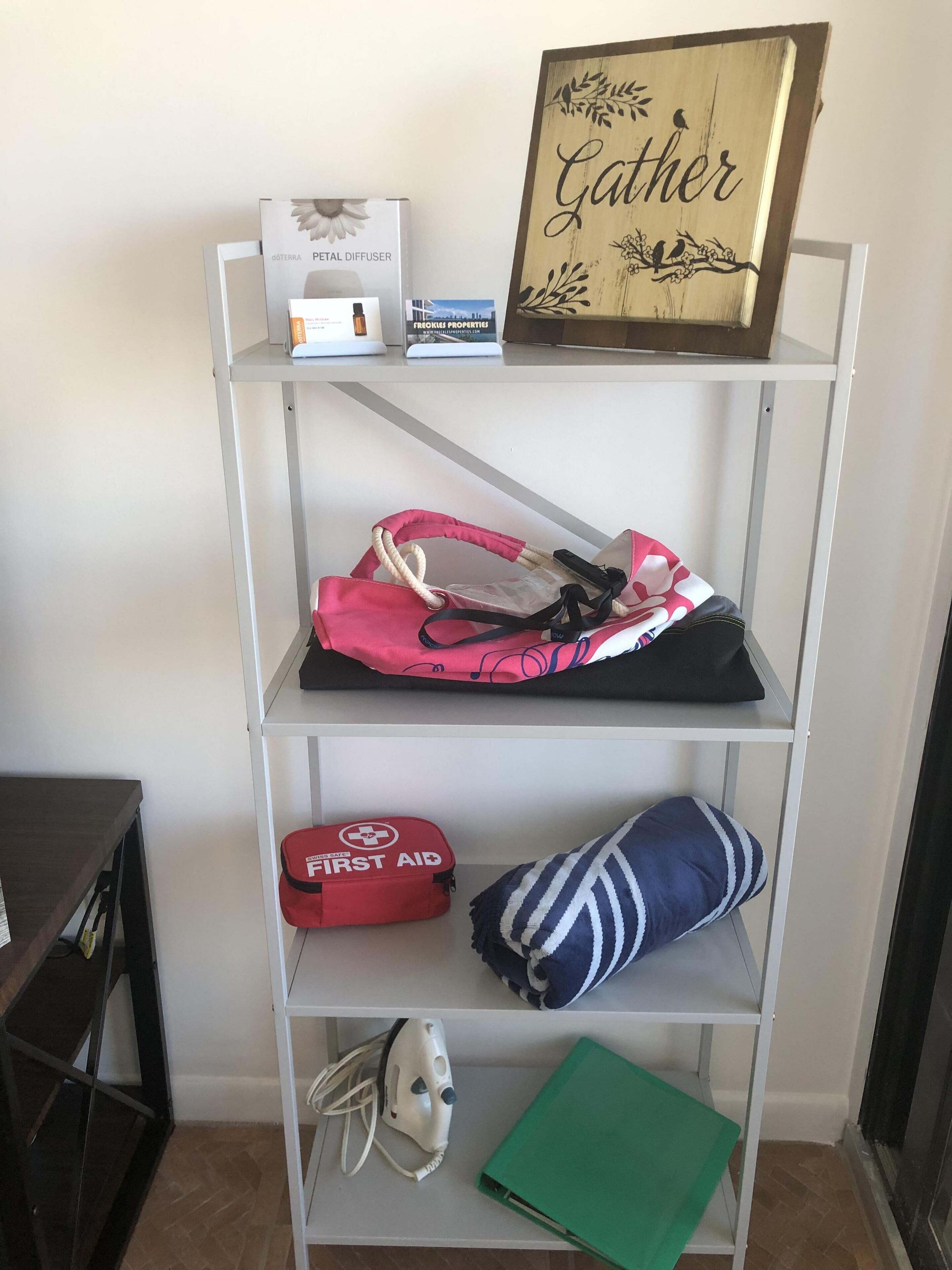 A shelf with a perfume diffuser, a red first aid kit, a blue and white striped pillow, a green folder, and a framed sign with the word "Gather".