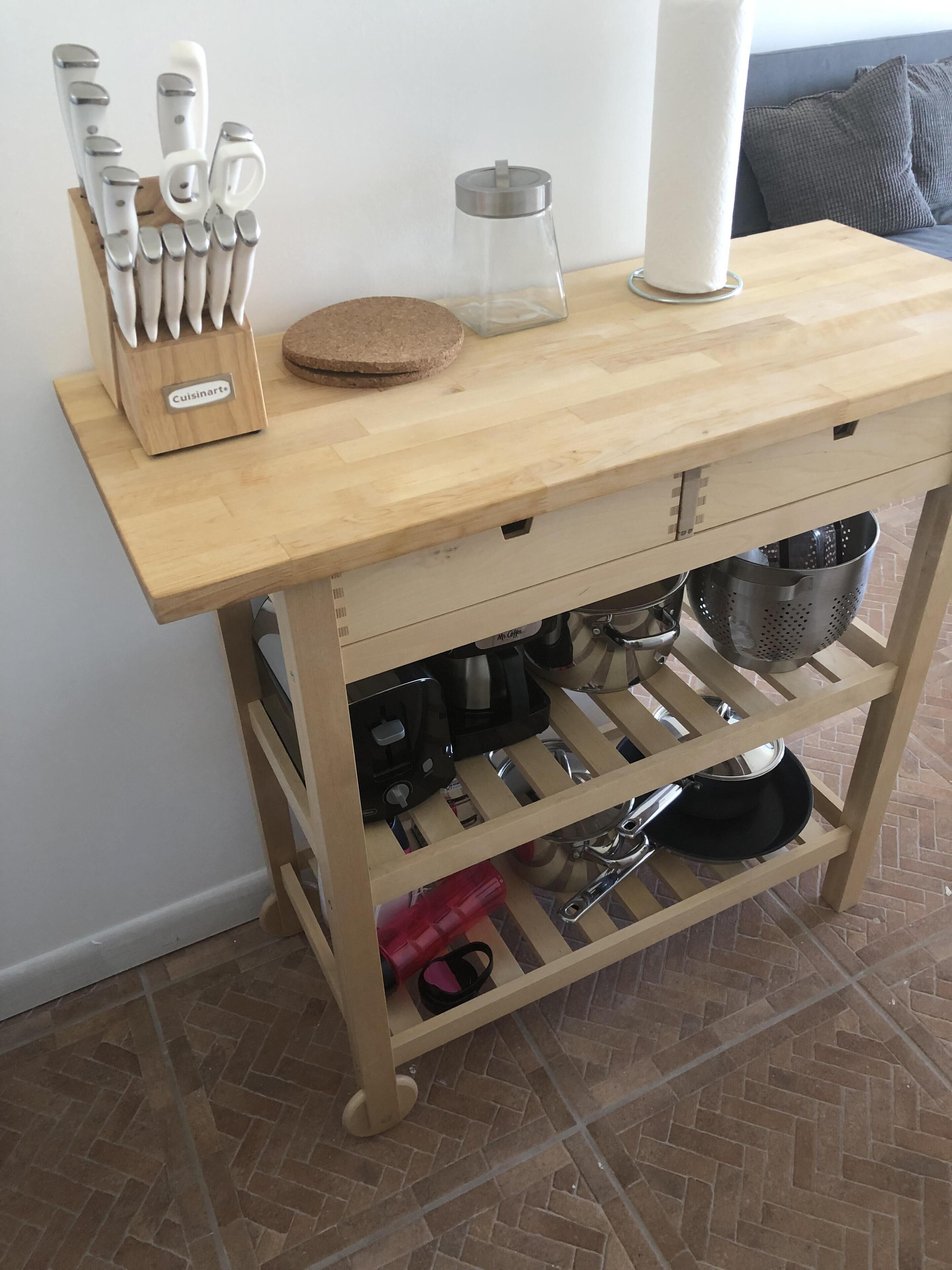 Un carrito de cocina de madera con tabla de cortar, un bloque para cuchillos, un colador y varios utensilios de cocina.