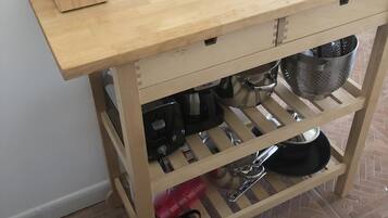 Un carrito de cocina de madera con tabla de cortar, un bloque para cuchillos, un colador y varios utensilios de cocina.